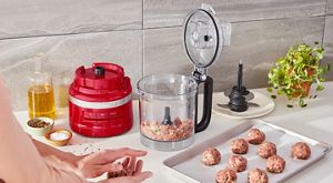 Open food processor with ground meat next to a tray of rolled ground meat and a hand rolling meatballs
