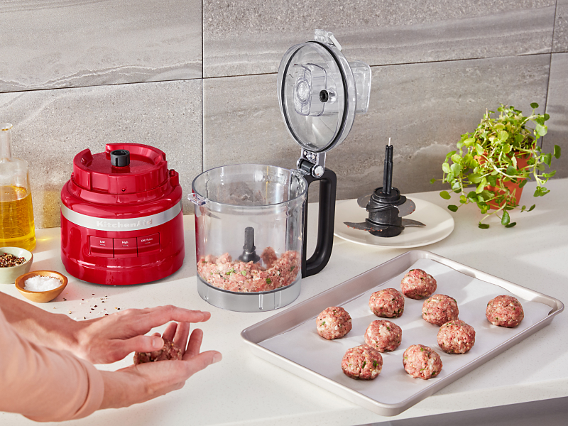 Open food processor with ground meat next to a tray of rolled ground meat and a hand rolling meatballs