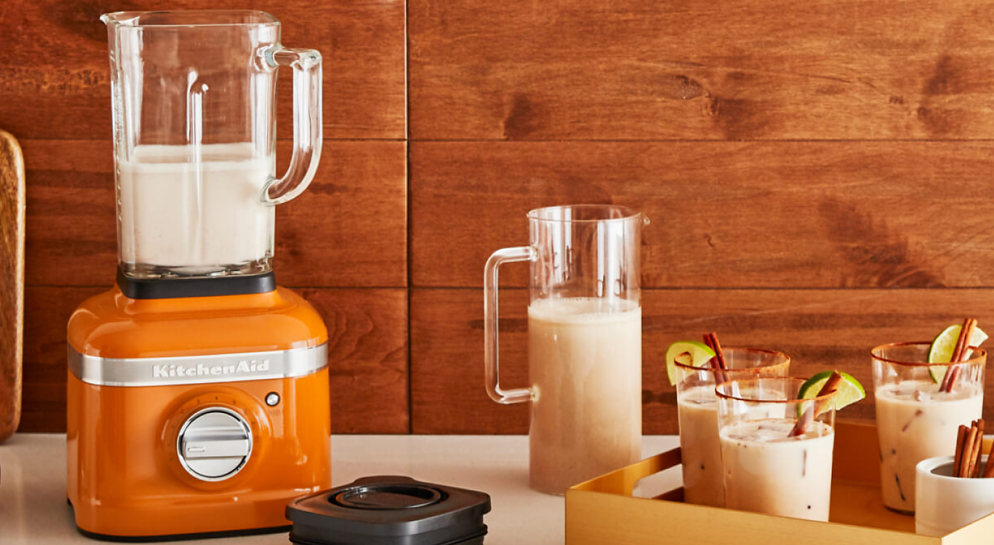 KitchenAid® Honey colored blender with white liquid sitting beside tray holding three cocktails garnished with limes and cinnamon sticks.