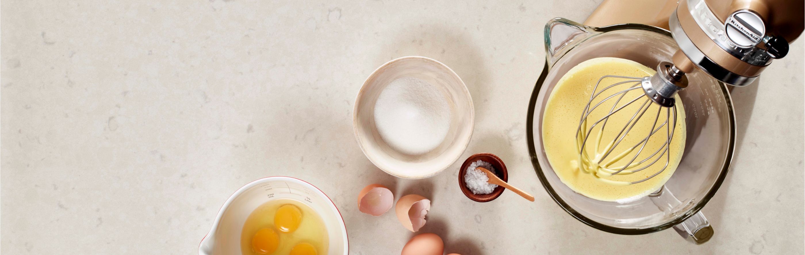 KitchenAid® stand mixer with whipped egg whites in the mixing bowl next to a bowl of cracked eggs and salt.