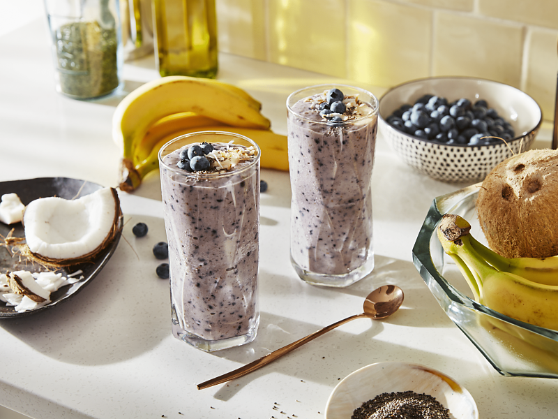 Two blueberry smoothies on countertop with bananas and coconut ingredients