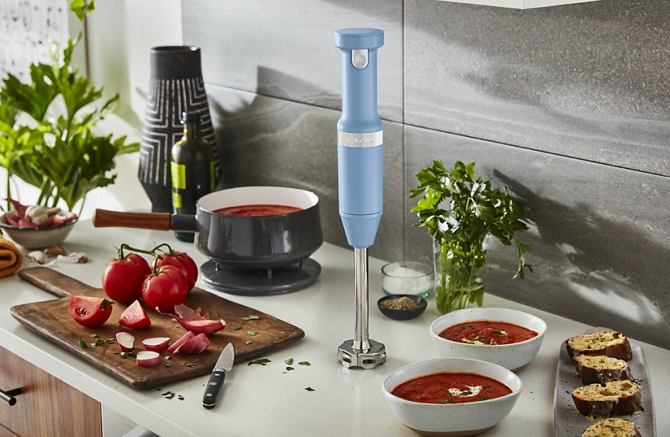 KitchenAid® hand blender in Blue Velvet next to pot of tomato sauce and fresh tomatoes