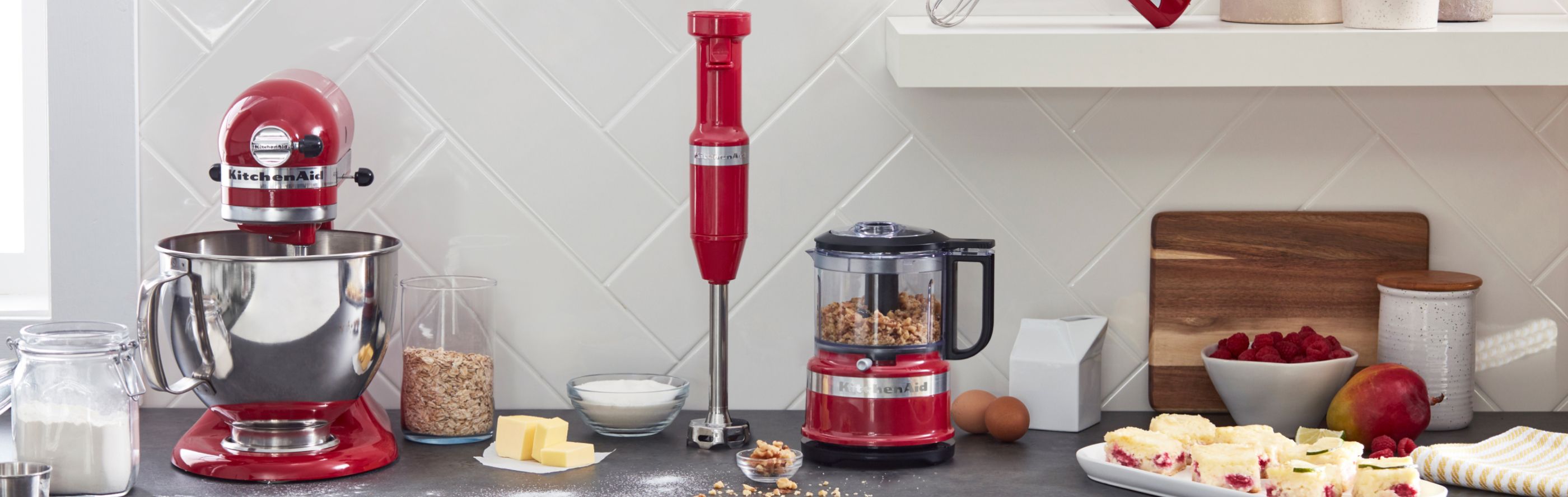 Red food processor next to red immersion blender from KitchenAid brand on counter with ingredients