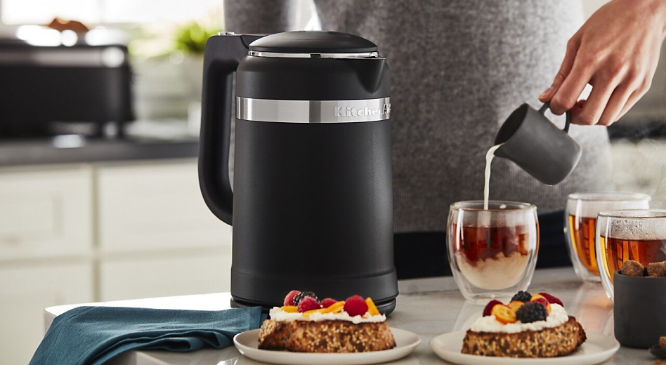 Hand pouring milk into cups of tea sitting next to electric tea kettle and plates of pastries. Hand pouring milk into cups of tea sitting next to electric tea kettle and plates of pastries.