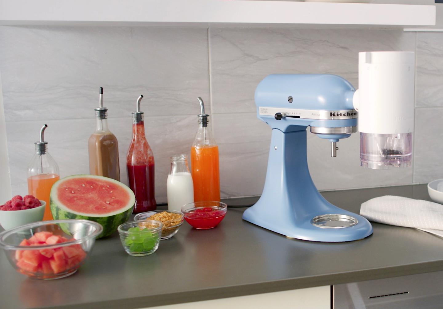 Blue stand mixer with KitchenAid® Shave Ice Attachment on counter with shaved ice syrups, watermelon and ingredients