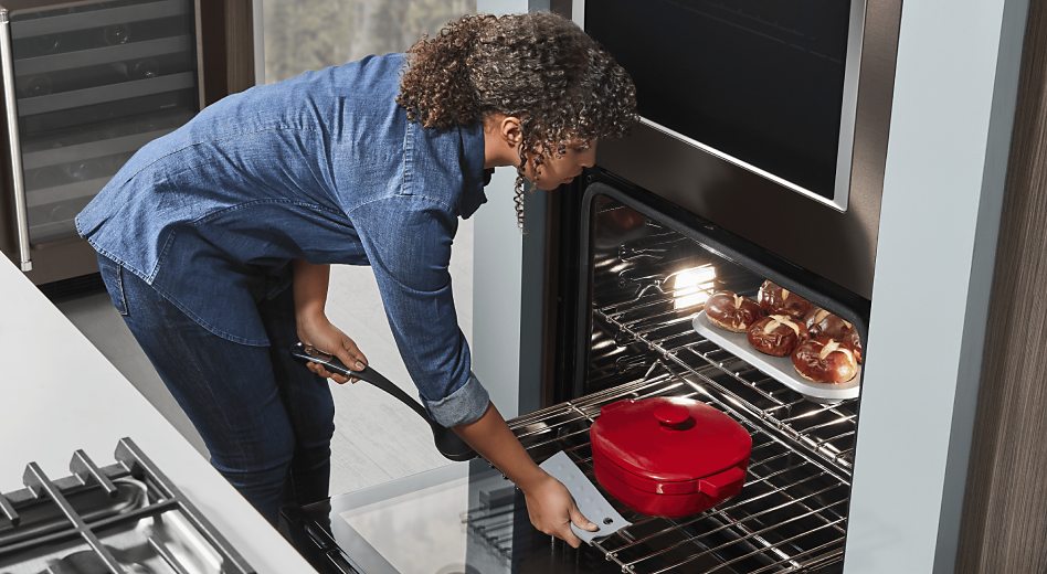 Woman placing red dutch oven in oven with homemade pretzel rolls baking on upper rack