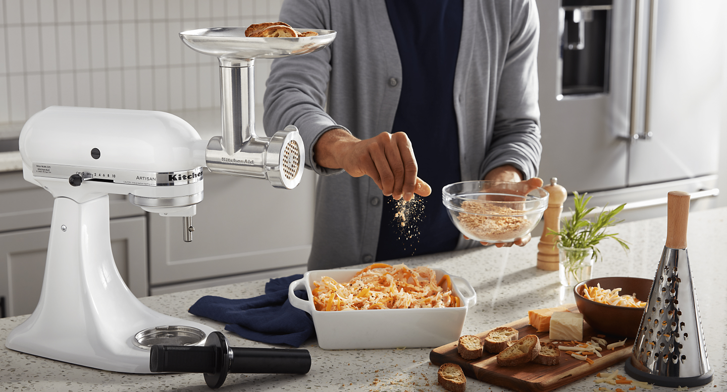 Male sprinkling bread crumbs in a dish next to a grater, wooden platter with bread and KitchenAid® stand mixer. 