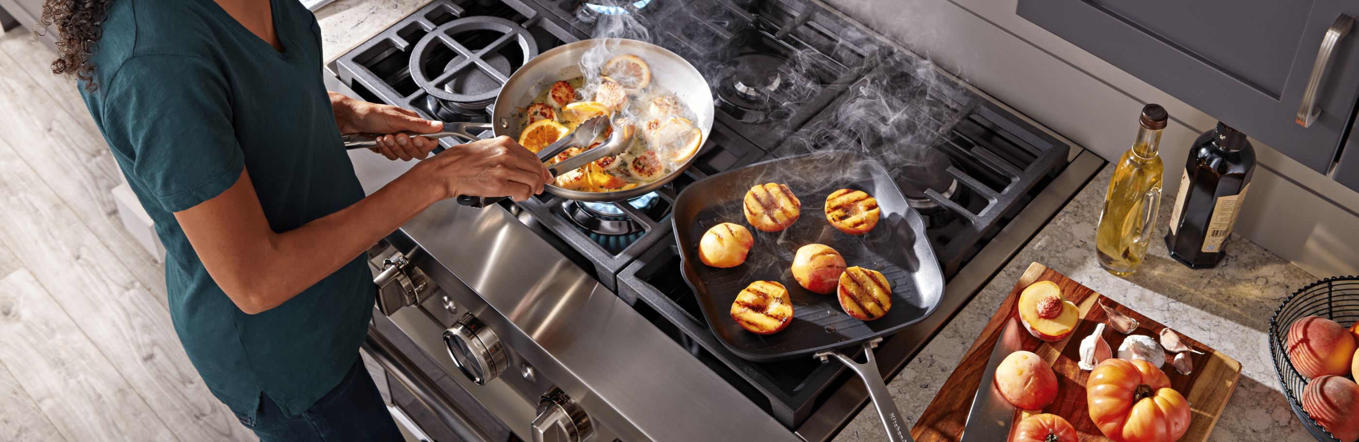 Female cooking scallops and citrus in a frying pan and grilling peaches in a grill pan on a gas range.
