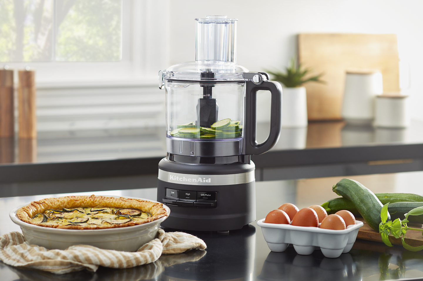 Sliced cucumbers in a black KitchenAid® food processor next to a vegetable quiche
