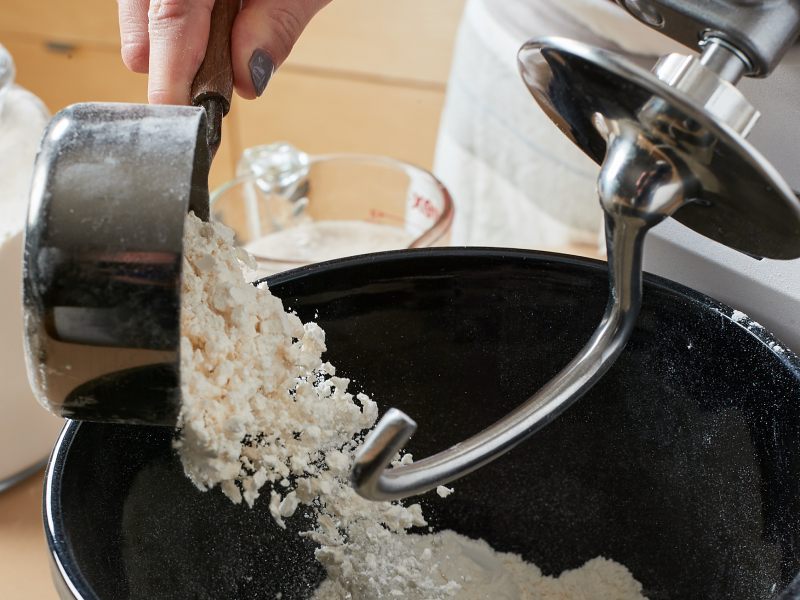 Person pouring flour into stand mixer equipped with a dough hook accessory