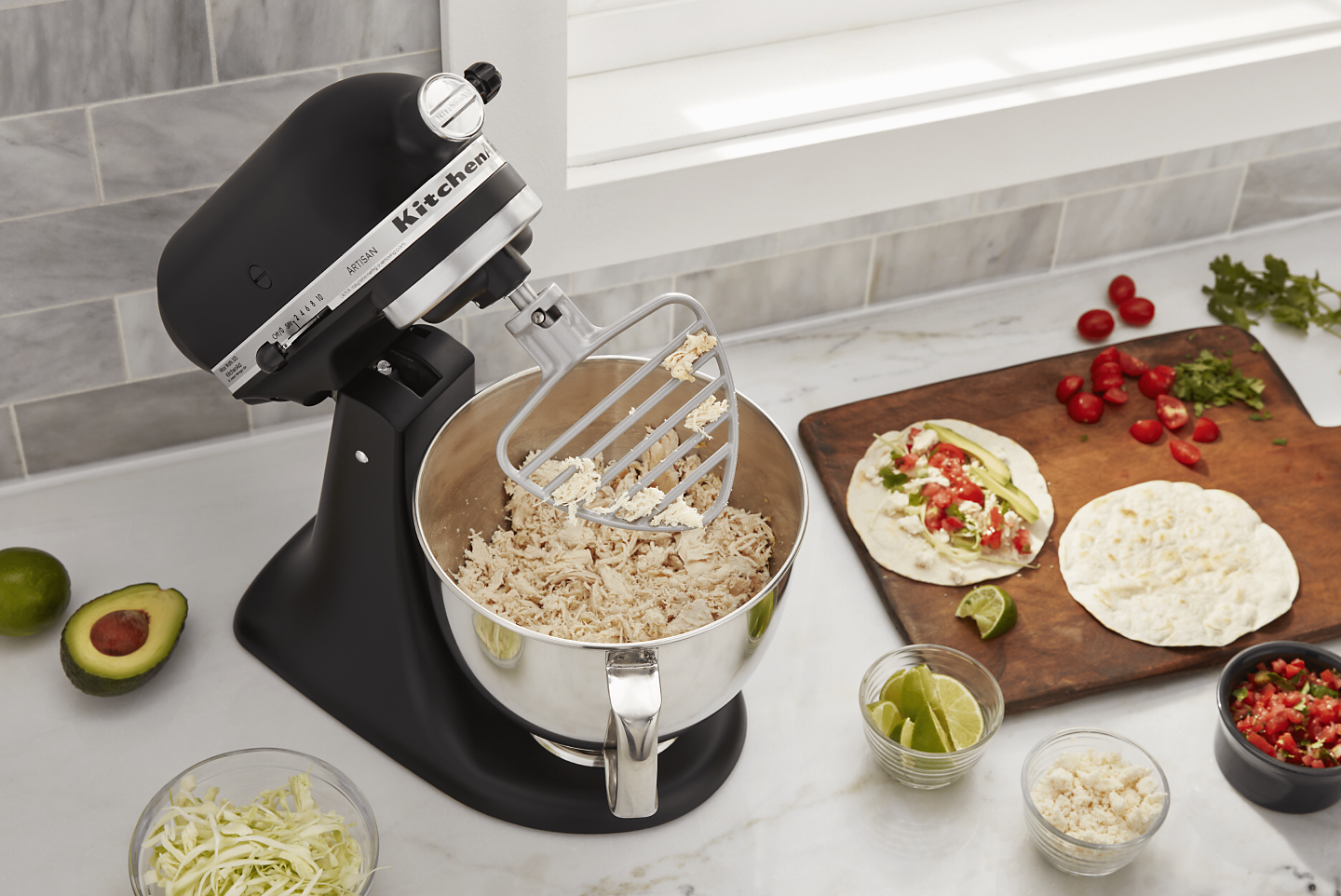 Black KitchenAid® Stand Mixer shredding chicken using the paddle attachment while surrounded by tortillas, salsa and taco fixings on a counter 