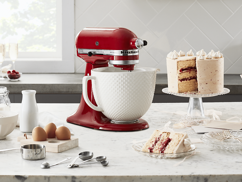 Red KitchenAid® Stand Mixer on a counter next to a three layer white frosted cake, milk, eggs, measuring cups and flour 