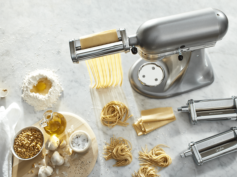 Silver KitchenAid® Stand Mixer with pasta-making attachments surrounded by mounds of fresh pasta and pasta ingredients 