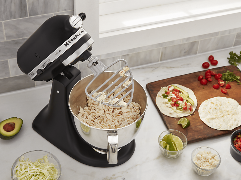 Black KitchenAid® Stand Mixer shredding chicken using the paddle attachment while surrounded by tortillas, salsa and taco fixings on a counter 