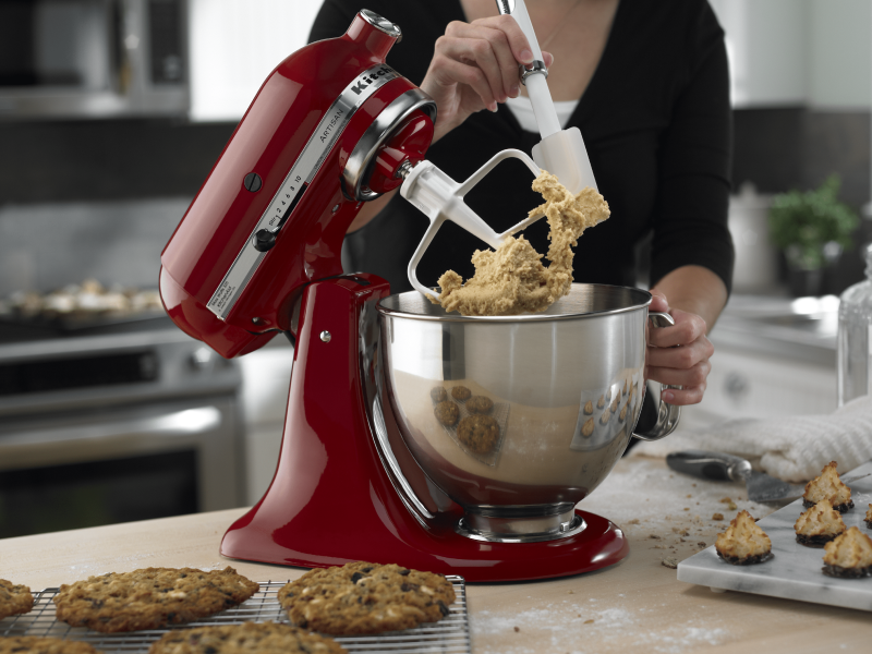 Tilt-head red KitchenAid® Stand Mixer with paddle attachment being scraped down while mixing up cookie dough with baked cookies in the foreground 