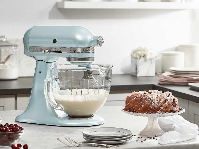 Pearly blue KitchenAid® Stand Mixer with glass bowl whipping cream next to a bundt cake sprinkled with confectioners sugar 