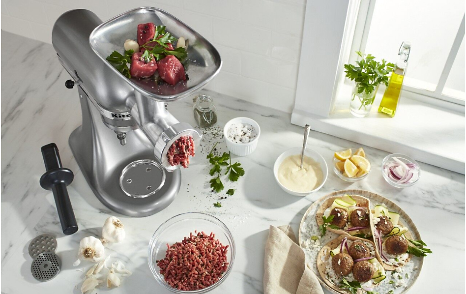 Silver stand mixer with food grinder attachment grinding meat surrounded by ingredients