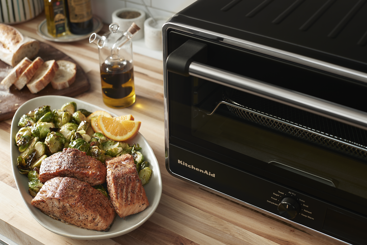 KitchenAid® countertop oven next to a plate of cooked salmon with roasted brussel sprouts and lemon wedges