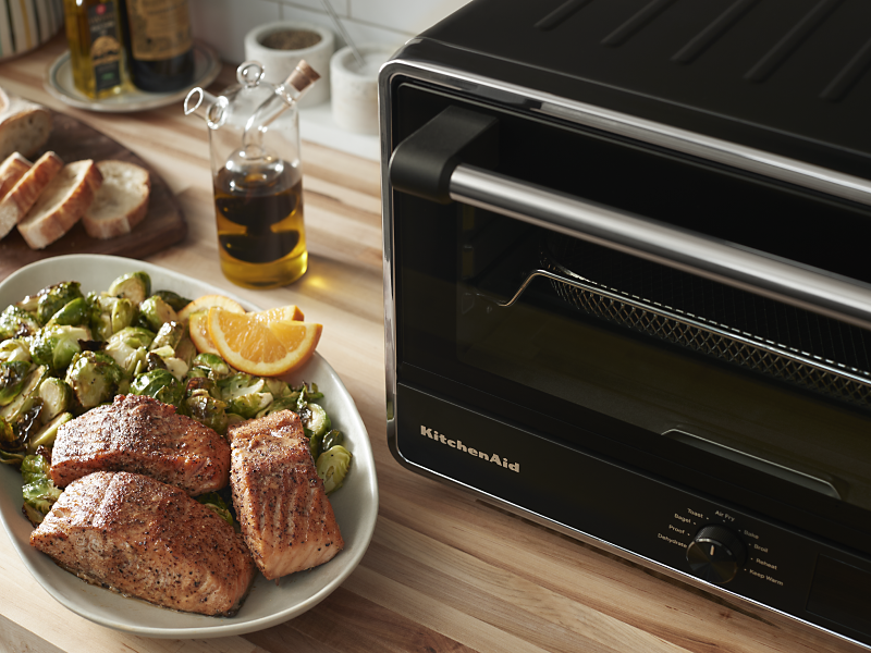 KitchenAid® countertop oven next to a plate of cooked salmon with roasted brussel sprouts and lemon wedges