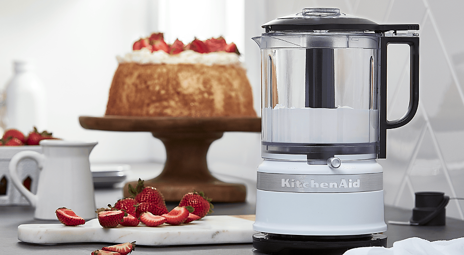 A KitchenAid® food processor in front of a cake topped with strawberries