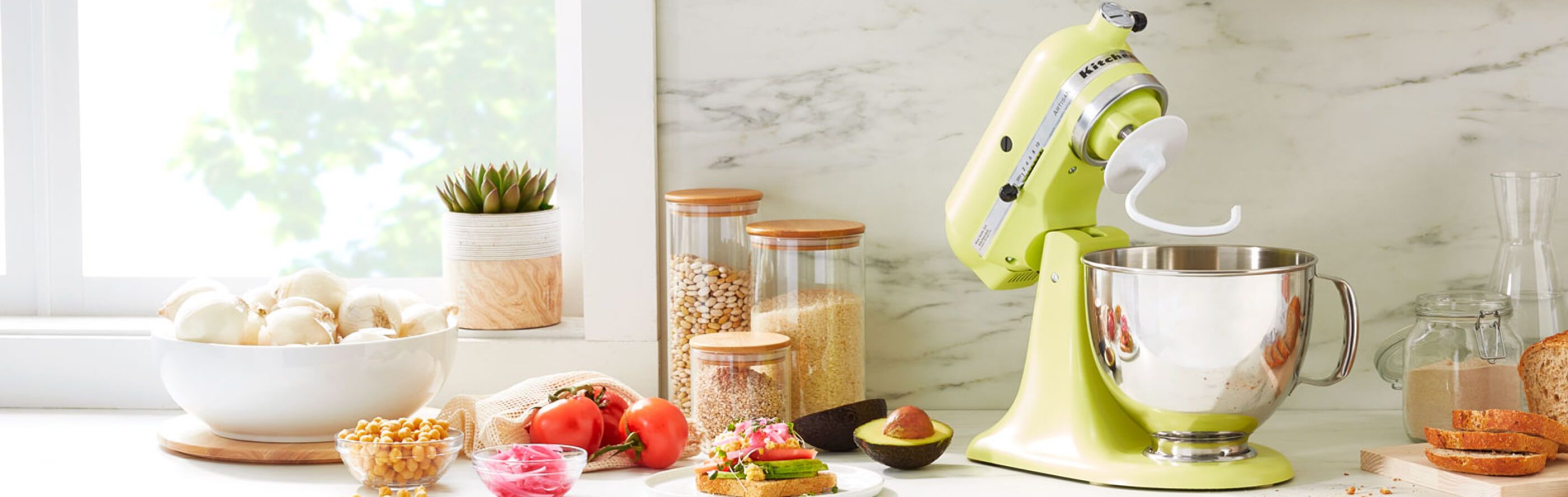 A KitchenAid® stand mixer with a dough hook accessory on a modern kitchen counter next to fresh vegetables for making a sandwich.