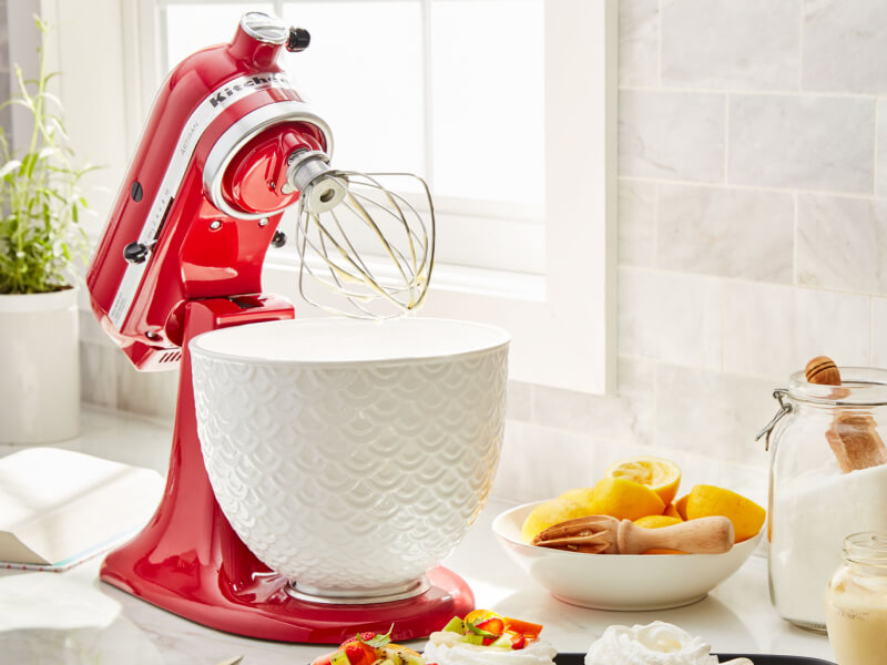 A KitchenAid® stand mixer with a wire whisk/whip accessory on the counter of a modern kitchen.