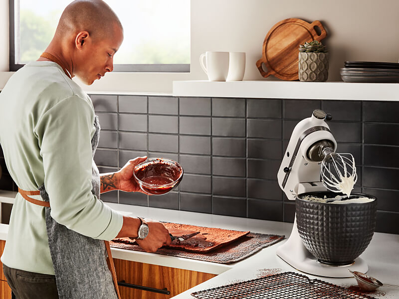A person with a bowl of melted chocolate standing next to a KitchenAid® stand mixer with a wire whisk accessory and whipped egg whites.