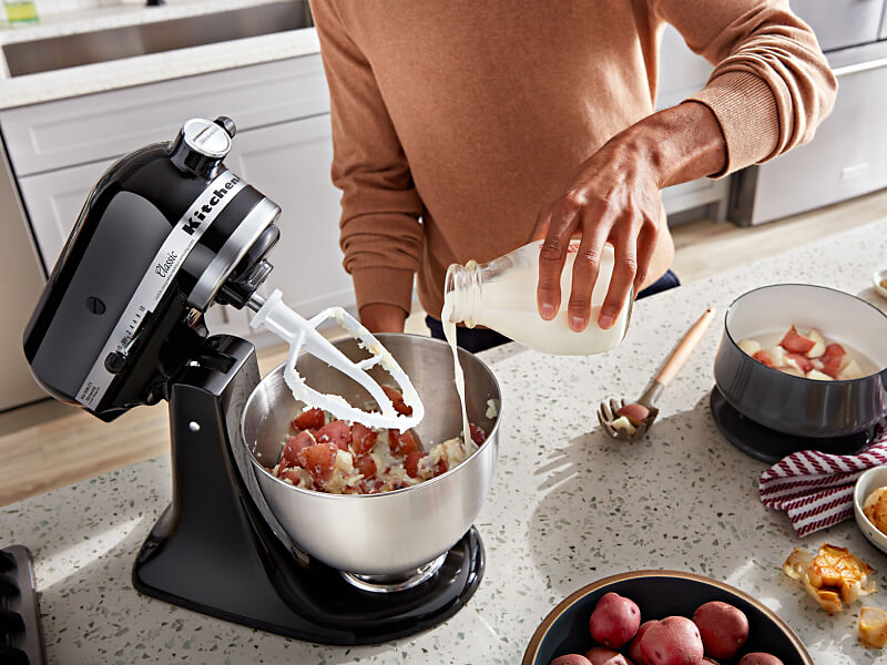 A person pouring milk into a KitchenAid® stand mixer bowl with ingredients and a flat beater accessory on the counter of a modern kitchen.