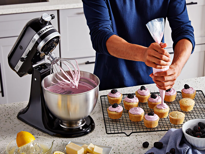 A person frosting cupcakes on the counter of a modern kitchen next to a KitchenAid® stand mixer with a wire whisk/whip accessory.