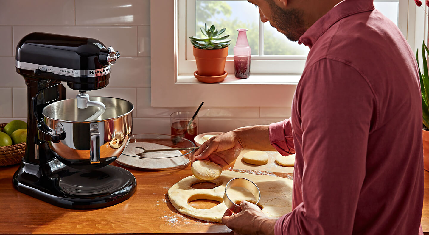 A person cutting biscuit dough on the counter of a modern kitchen next to a KitchenAid® stand mixer with a dough hook accessory.
