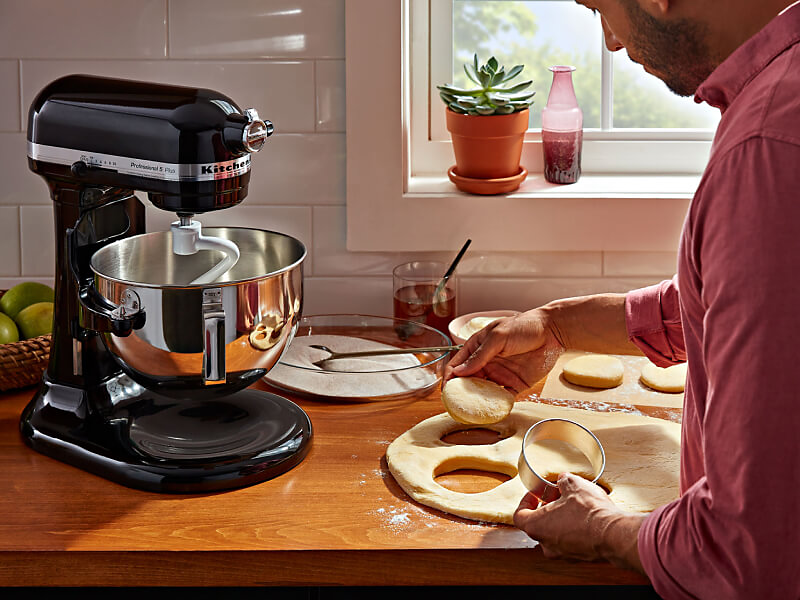 A person cutting biscuit dough on the counter of a modern kitchen next to a KitchenAid® stand mixer with a dough hook accessory.