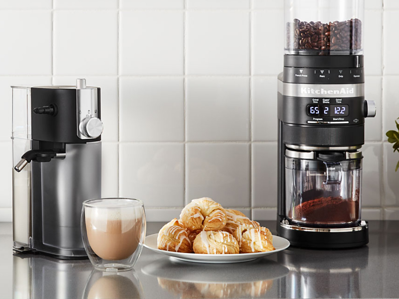 A cup of whipped coffee on a stainless steel countertop with baked goods and appliances