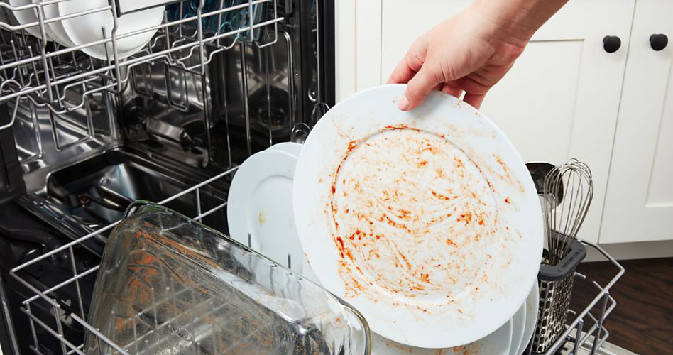 Someone holding a dirty plate over a Maytag dishwasher. On the racks of the dishwasher are plates, utensils, bowls and a baking dish