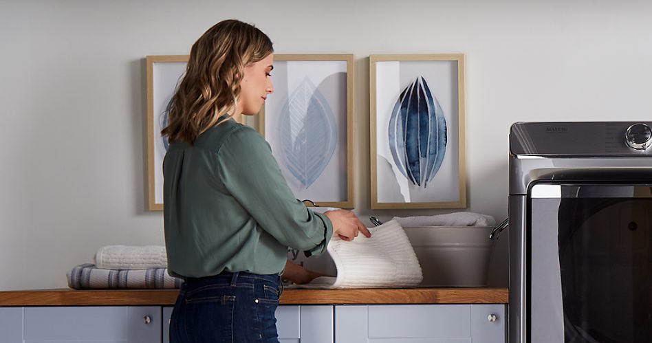 A woman wearing a green blouse and jeans in her laundry room. She is folding towels. Behind her, three framed pictures hang on the wall and there is a counter above cabinets. Next to her is a Maytag washing machine.