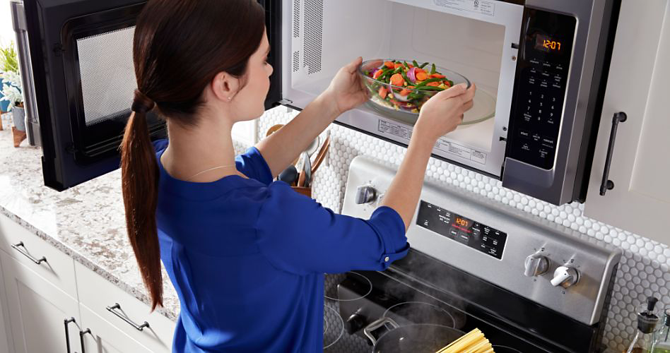 A woman places a bowl of vegetables in an Maytag over-the-range microwave. Below is a Maytag oven with pasta cooking in a pot of boiling water on a burner