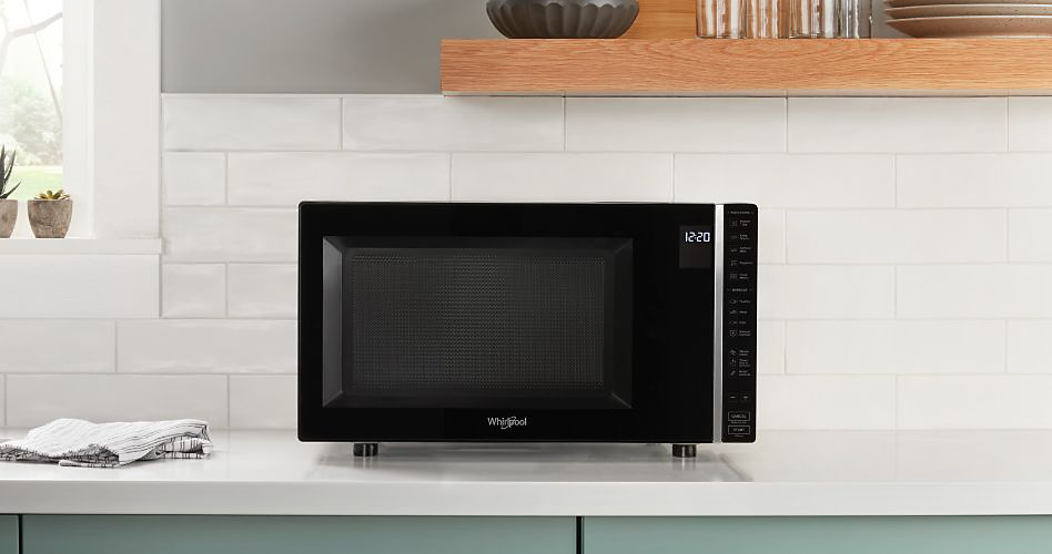 A Maytag countertop microwave. On the counter next to it is a dish cloth. There are cacti on the window sill behind it. Above is a shelf with a stack of plates, glasses and a ceramic bowl