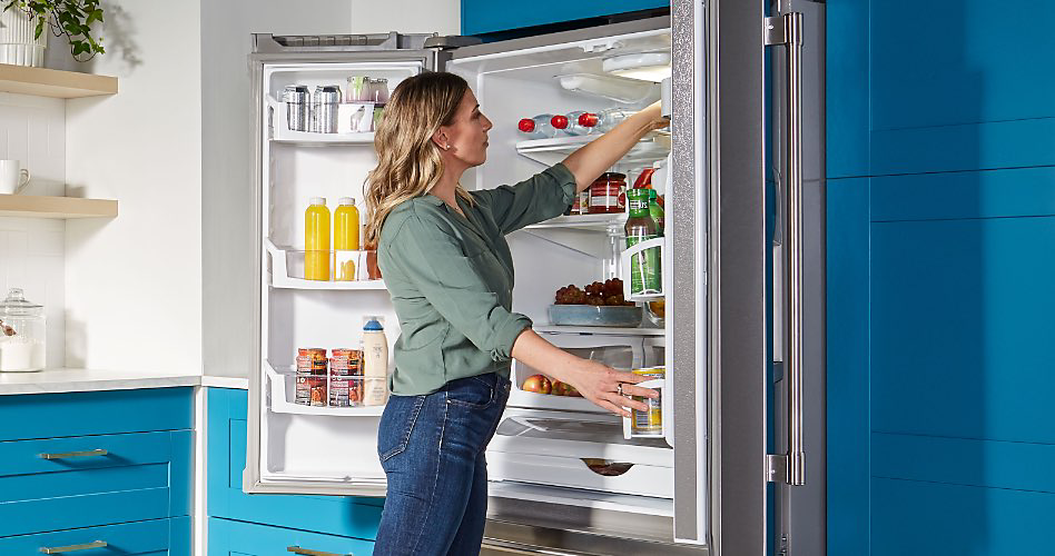 alt="A woman in front of an opened refrigerator. Inside the fridge are beverage bottles, condiments, bottles of dressing, water bottles and more, and she is surrounded by blue drawers and cabinets."