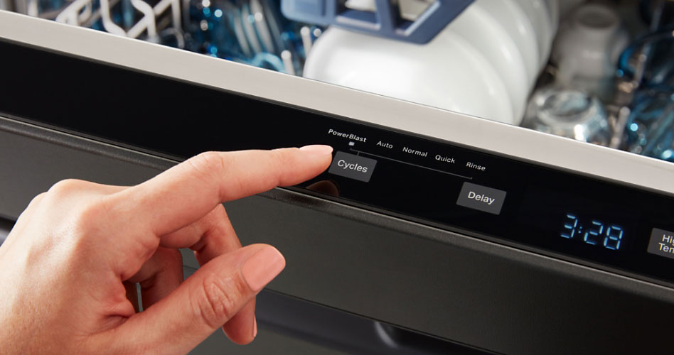 Someone pressing a button on the top control of a Maytag fully-integrated dishwasher. Inside on the top rack are bowls and glasses.