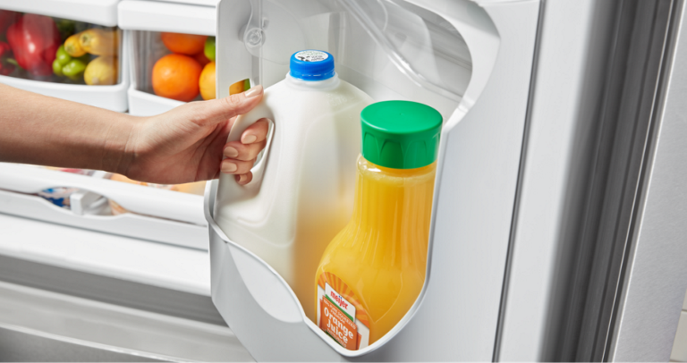 An opened Maytag Refrigerator. Someone is holding the handle of a jug of milk. Next to the milk is orange juice. Also in the fridge are various produce. 