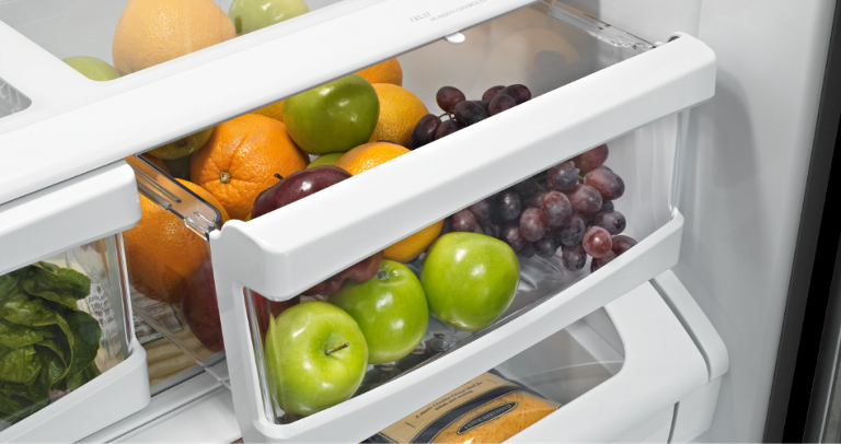   Inside a Maytag Refrigerator. The crisper drawer is slightly opened and inside are green apples, oranges and red grapes.
