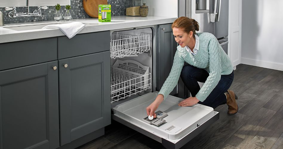 Someone places an affresh pod in the detergent compartment in their Maytag dishwasher. A package of affresh cleaner is on the counter next to the sink. The kitchen has a Maytag fridge and grey cabinets.