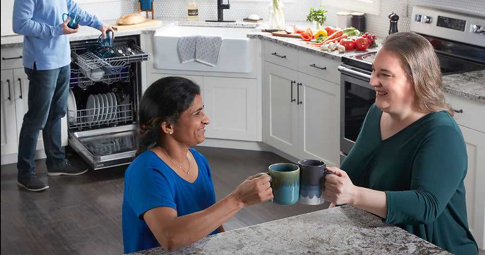 Two women cheer their coffee mugs at a kitchen island. In the background are a Maytag Oven, a counter full of produce, a sink and a man loading a Maytag Dishwasher.