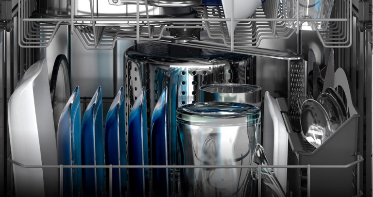 The interior of a Maytag Dishwasher. The racks are filled with corning ware, plates, mugs, a full cutlery tray and a colander pot.