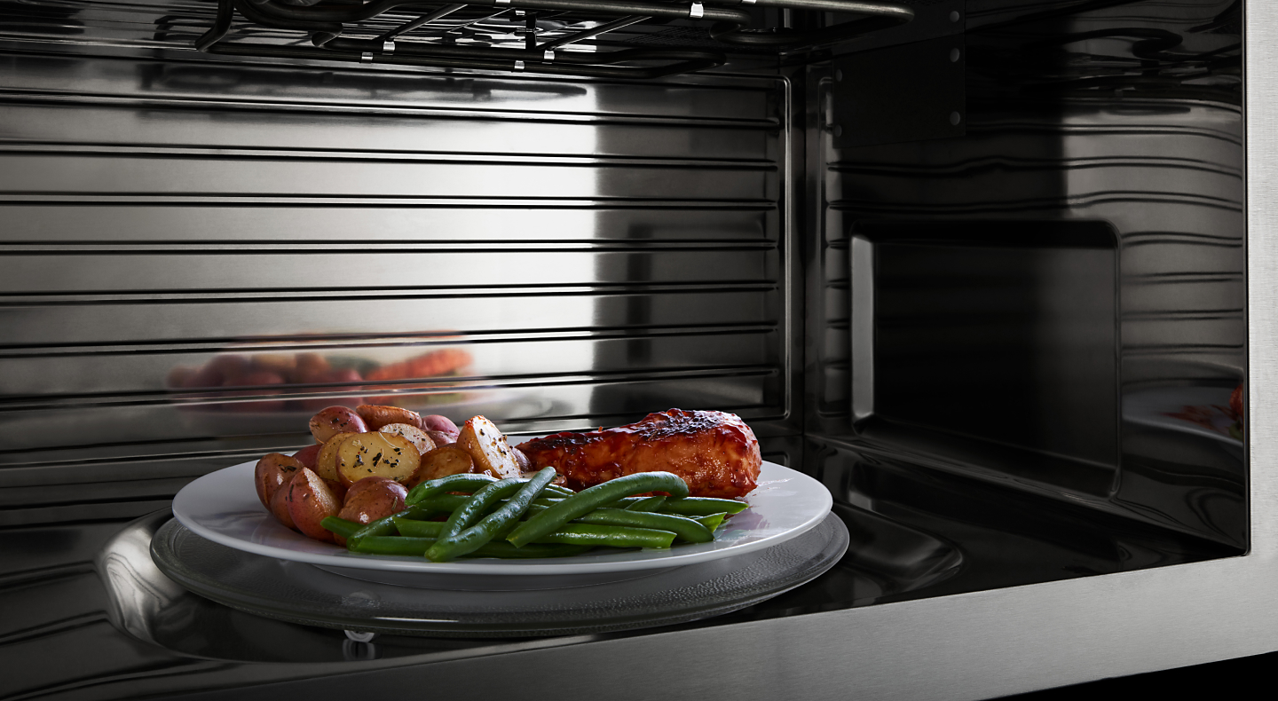 A closeup of the interior of a convection Maytag® microwave heating chicken, potatoes and beans.