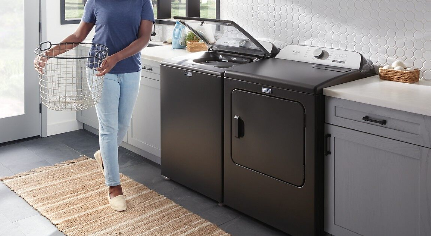 Person holding empty laundry basket next to open top load Maytag® washer and dryer pair.
