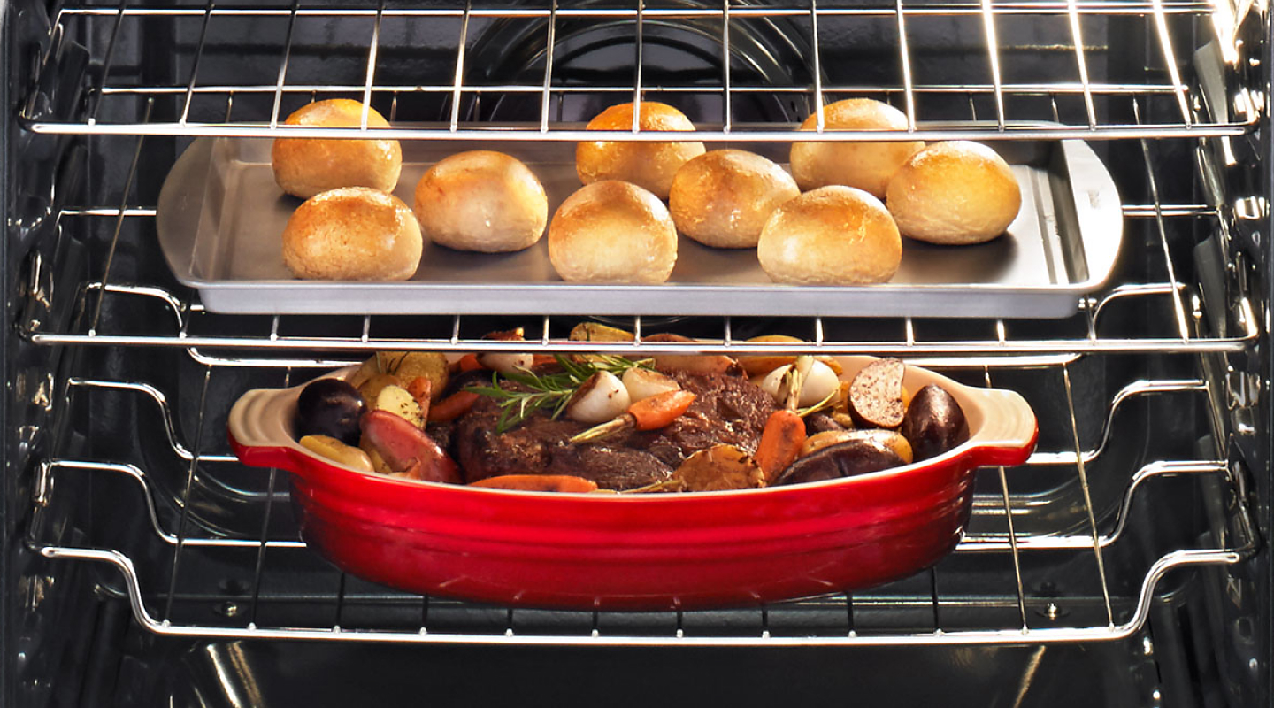 Meat with roasted vegetables cooking under a baking sheet with rolls
