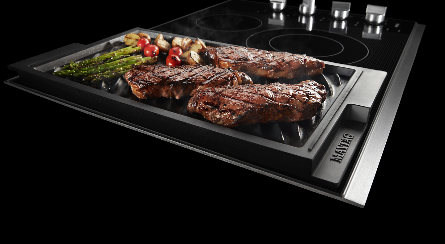 Steaks and vegetables cooking in a cast iron grill pan on a Maytag® electric cooktop