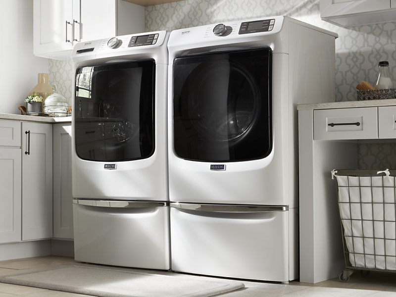 Side-by-side white Maytag® front load washer and dryer on pedestals in laundry room