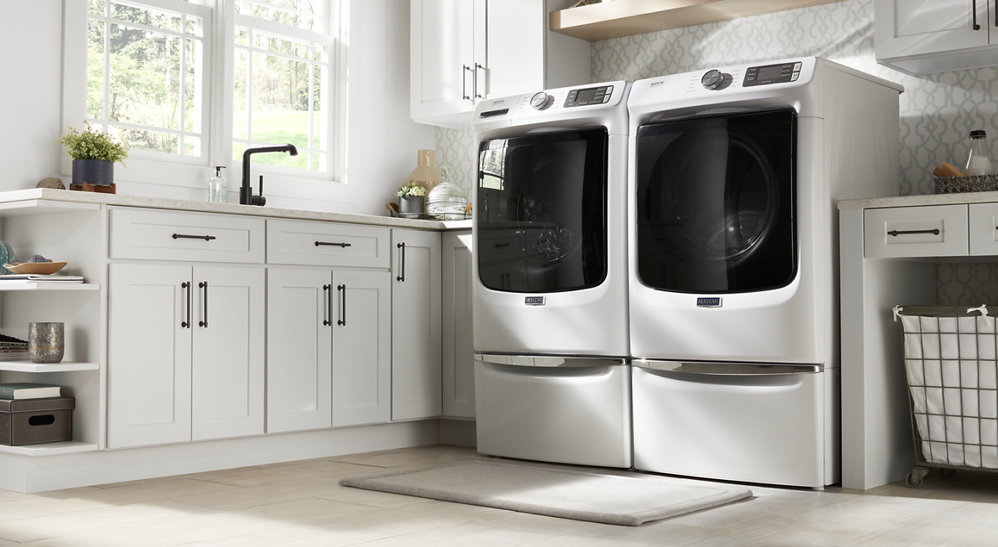 Side-by-side white Maytag® front load washer and dryer on pedestals in laundry room