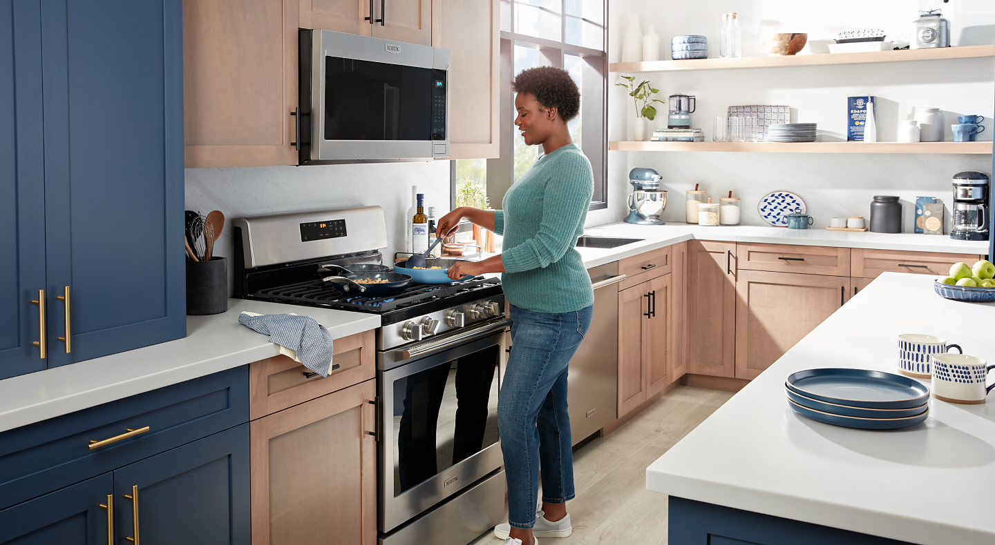 A person sautéing food on the cooktop of a Maytag® gas oven with an over-the-range microwave in a modern kitchen